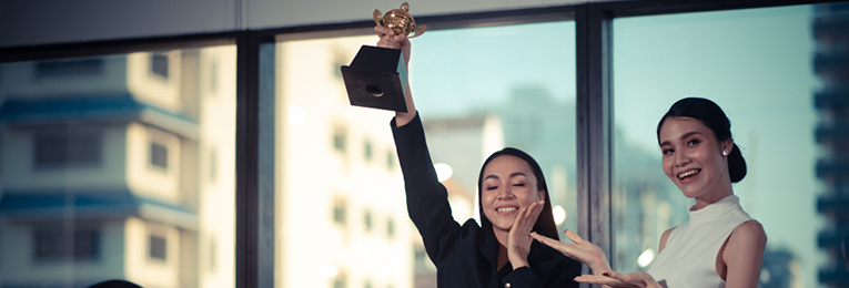 A woman proudly raises a trophy in the air as another woman points to her, smiling and looking at the camera.